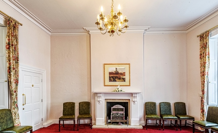 A room with a carpet floor and some chairs at the back. A fireplace is in the middle of the back wall and has some art above it. A chandelier hangs from the ceiling.